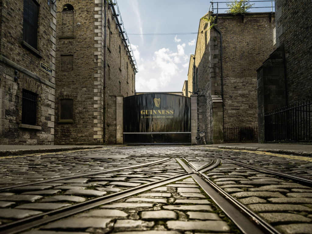 De geplaveide straat leidt naar een grote zwarte poort met het Guinness-logo en het gouden harpembleem, perfect voor degenen die een rondreis Ierland maken. Omringd door hoge, oude bakstenen gebouwen onder lichte wolken en een blauwe lucht, benadrukt zonlicht de historische ambiance met tramrails die de voorgrond kruisen. | Echt Ierland Vakanties
