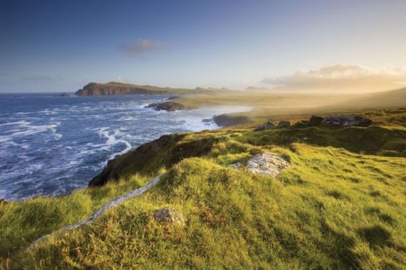 Ervaar de betoverende aantrekkingskracht van een vakantie in Ierland met een kustlandschap bij zonsondergang, met grasachtige kliffen op de voorgrond. Golven beuken tegen de rotsachtige kust onder een heldere hemel, terwijl gouden licht de verre heuvels verlicht, waardoor een serene en natuurlijke omgeving ontstaat die perfect is voor uw Ierse reis. | Echt Ierland Vakanties