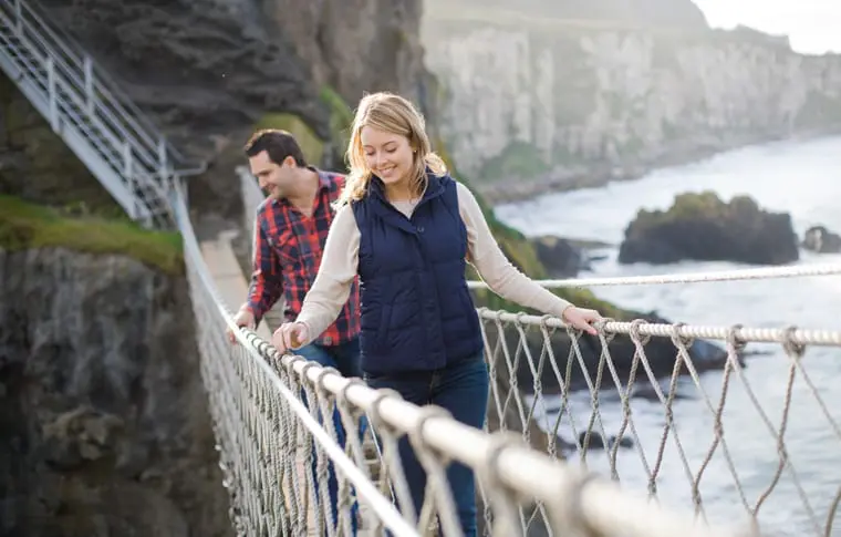 Een vrouw en een man lopen over een touwbrug in Ierland. De vrouw, in een marineblauw vest, staat op de voorgrond lachend, terwijl de man in een rood geruit overhemd volgt. Op de achtergrond zijn kliffen en een watermassa onder een bewolkte lucht te zien.