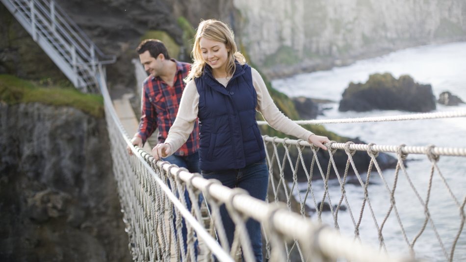 Een vrouw en een man lopen over een touwbrug in Ierland. De vrouw, in een marineblauw vest, staat op de voorgrond lachend, terwijl de man in een rood geruit overhemd volgt. Op de achtergrond zijn kliffen en een watermassa onder een bewolkte lucht te zien.
