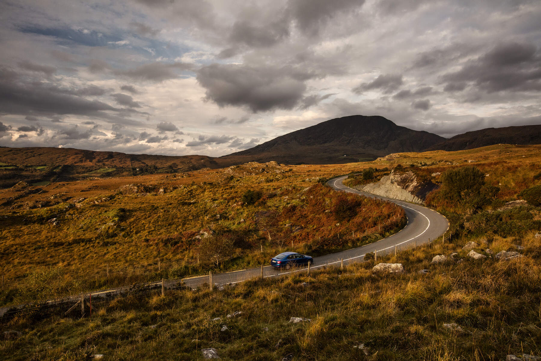 Een kronkelende weg slingert door een uitgestrekt landschap van glooiende heuvels en rotsachtig terrein onder een bewolkte lucht, perfect voor een onvergetelijke vakantie in Ierland. Een blauwe auto rijdt over de weg. Op de achtergrond domineert een grote berg het tafereel, met stukken groen verspreid over het terrein. De verlichting is warm en goudkleurig. | Echt Ierland Vakanties