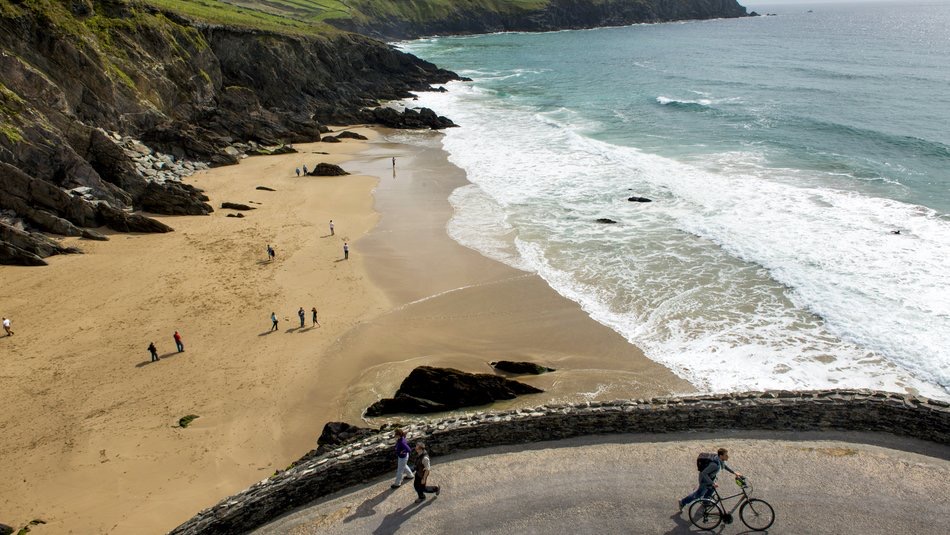 Een schilderachtig uitzicht op de kust toont een kronkelend pad langs een stenen muur boven een zandstrand, perfect voor degenen die op zoek zijn naar een vakantie in Ierland. Mensen slenteren langs de kust terwijl golven zachtjes tegen het zand klotsen, terwijl een fietser geniet van het pad. Weelderige groene kliffen rijzen op de achtergrond op en ontmoeten de blauwe zee. | Echt Ierland Vakanties