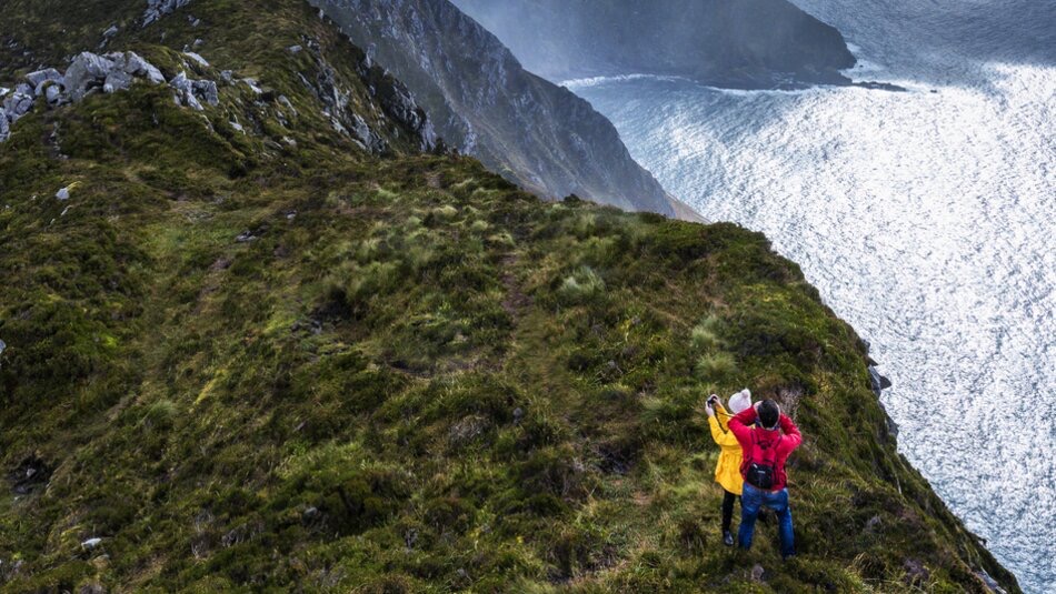 Twee mensen staan op een met gras begroeide klifrand in Ierland en leggen het moment vast met een selfie. De een draagt een rood jasje, de ander een geel. De zee glinstert onder de bewolkte lucht en ruige kliffen vormen een prachtig decor voor hun vakantie in Ierland. | Echt Ierland Vakanties