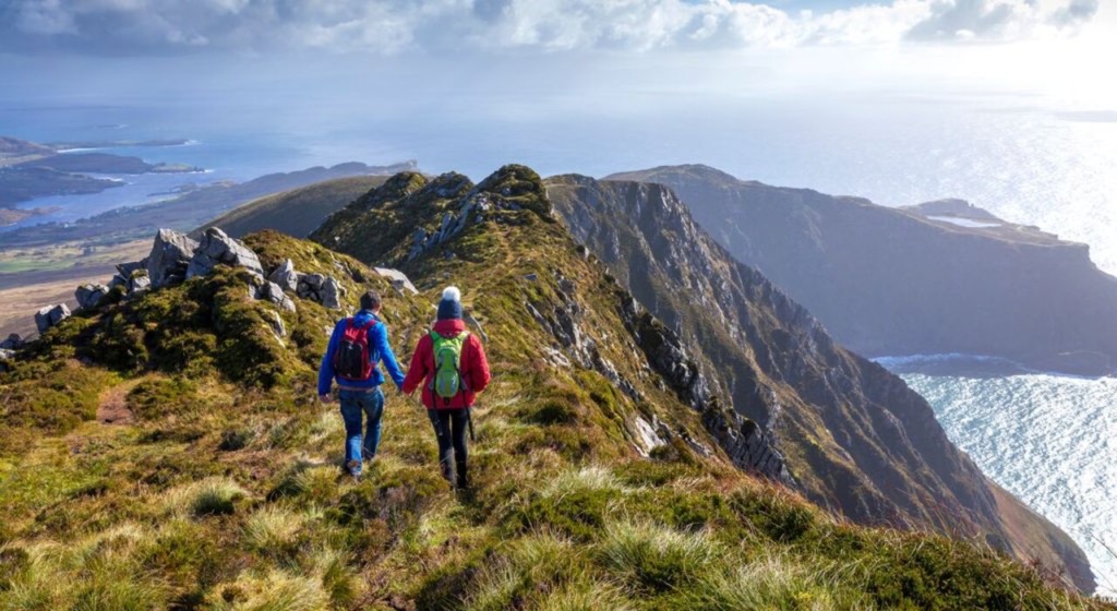 Twee wandelaars trekken over een met gras begroeide bergrug met uitzicht op de uitgestrekte oceaan, wat de essentie van een vakantie Ierland belichaamt. Met kleurrijke rugzakken en jassen genieten ze van de gedeeltelijk bewolkte luchten die een mix van zonlicht en schaduw werpen over de ruige kliffen die dit prachtige Ierse landschap definiëren. | Echt Ierland Vakanties