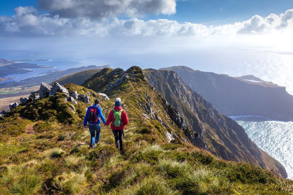Twee wandelaars in kleurrijke jassen lopen over een met gras begroeide bergkam richting rotsachtige kliffen met uitzicht op een uitgestrekte oceaan, perfect voor degenen die een inspirerende rondreis Ierland maken. De lucht is gedeeltelijk bewolkt, zonlicht weerkaatst op het water en weelderige landschappen met verre eilanden maken deze onvergetelijke vakantie Ierland-ervaring compleet. | Echt Ierland Vakanties