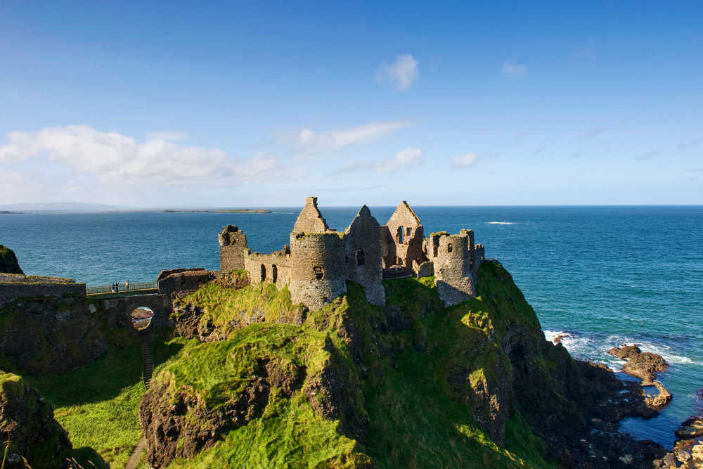 Luchtfoto van Dunluce Castle ruïnes, gelegen op een rotsachtige klif met uitzicht op de oceaan in Noord-Ierland, perfect voor degenen die een onvergetelijke vakantie in Ierland plannen. De stenen structuur is omgeven door weelderig groen gras. Een voetgangersbrug verbindt de overblijfselen met het vasteland onder een helderblauwe lucht. | Echt Ierland Vakanties