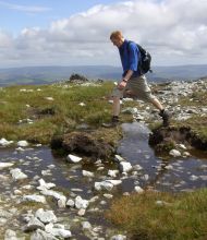 Een persoon in wandelkleding springt over een rotsachtige stroom in een grasachtig berglandschap tijdens hun vakantie in Ierland. Ze dragen een blauw shirt met lange mouwen, een beige broek en bruine laarzen, met een zwarte rugzak. De lucht is gedeeltelijk bewolkt en op de achtergrond zijn verre heuvels zichtbaar. | Echt Ierland Vakanties