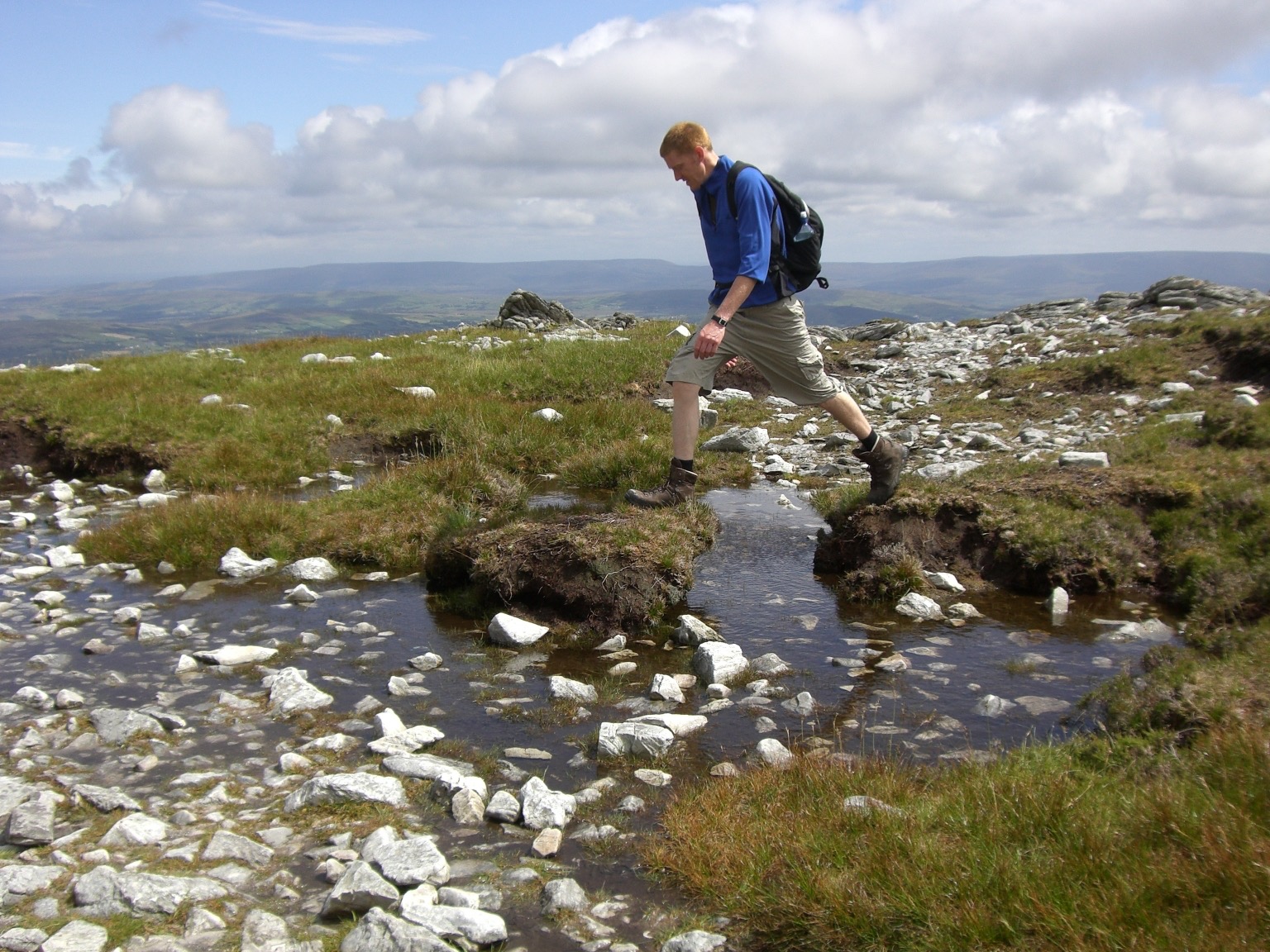 Een persoon in wandelkleding springt over een rotsachtige stroom in een grasachtig berglandschap tijdens hun vakantie in Ierland. Ze dragen een blauw shirt met lange mouwen, een beige broek en bruine laarzen, met een zwarte rugzak. De lucht is gedeeltelijk bewolkt en op de achtergrond zijn verre heuvels zichtbaar. | Echt Ierland Vakanties