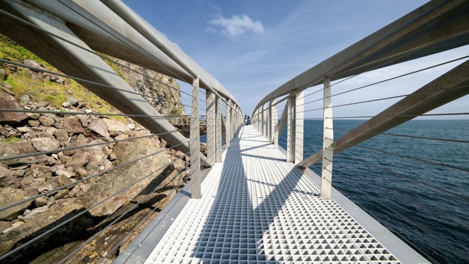 Een perspectiefweergave van een moderne voetgangersbrug met metalen leuningen en een geribbeld wandelpad dat zich uitstrekt over een rotsachtige kustlijn, wat doet denken aan een onvergetelijke rondreis Ierland. De brug leidt naar een helderblauwe lucht en kalme zee, met verre figuren zichtbaar aan het uiteinde. Rotsachtige kliffen zijn zichtbaar aan de linkerkant. | Echt Ierland Vakanties