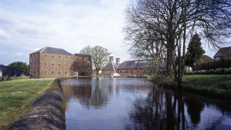 Een serene distilleerderijscène in het hart van een idyllische rondreis Ierland met een reflecterende vijver omzoomd door weelderig gras en bomen. De gebouwen met rode bakstenen accenten en opvallende dakopeningen staan majestueus, terwijl een gedeeltelijk bewolkte lucht de rustige sfeer versterkt. | Echt Ierland Vakanties