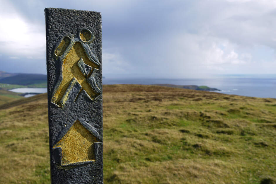 Close-up van een verweerde wandelroutemarkering met een gele figuur en pijl erin gekerfd, tegen een heuvelachtig landschap in het hart van Ierland. De achtergrond bestaat uit glooiende, met gras begroeide heuvels en de verre oceaan onder een gedeeltelijk bewolkte lucht, wat een avontuurlijke rondreis Ierland-ervaring belichaamt. | Echt Ierland Vakanties