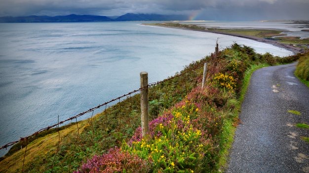 Ontdek dit kustlandschap dat doet denken aan vakantie Ierland, met een smal pad langs een klif omzoomd door weelderig gras en wilde bloemen. Een roestig hekwerk markeert de weg terwijl de uitgestrekte oceaan zich uitstrekt tot aan de horizon. Verre bergen onder een bewolkte lucht omlijsten een vage regenboog boven de rotsachtige kustlijn. | Echt Ierland Vakanties