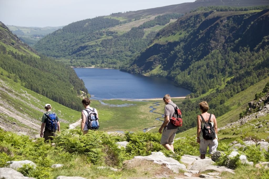 Vier wandelaars met rugzakken lopen over een rotsachtig pad naar een sereen meer, genesteld in een weelderige, groene vallei. Het landschap, dat doet denken aan een idyllische vakantie in Ierland, wordt omlijst door glooiende heuvels en dicht bos onder een helderblauwe lucht, wat een pittoresk en vredig tafereel oplevert. | Echt Ierland Vakanties