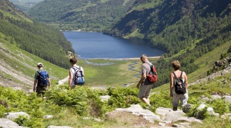 Vier wandelaars, met rugzakken, lopen over een rotsachtig pad naar een rustig meer omgeven door weelderige, groene heuvels in het hart van hun rondreis Ierland. Het meer ligt in een vallei met een lappendeken van bomen en vegetatie, onder een heldere hemel die het levendige daglicht reflecteert voor perfecte vakantie Ierland momenten. | Echt Ierland Vakanties