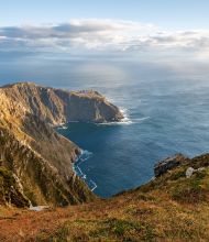 Een persoon in een groen jasje staat op een met gras begroeide klifrand, genietend van de schoonheid van hun vakantie in Ierland. Het uitzicht biedt een ruige kustlijn met dramatische kliffen en een kleine baai. De gedeeltelijk bewolkte lucht laat zonlicht delen van dit betoverende Ierse landschap verlichten. | Echt Ierland Vakanties