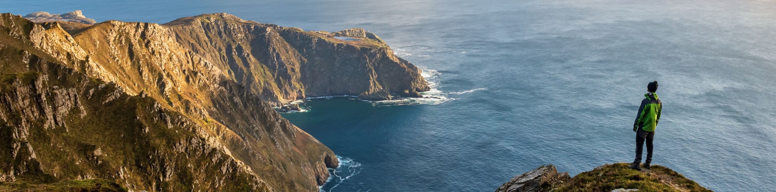 Een persoon in een groen jasje staat op een met gras begroeide klifrand, genietend van de schoonheid van hun vakantie in Ierland. Het uitzicht biedt een ruige kustlijn met dramatische kliffen en een kleine baai. De gedeeltelijk bewolkte lucht laat zonlicht delen van dit betoverende Ierse landschap verlichten. | Echt Ierland Vakanties