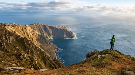 Een persoon in een groen jasje staat op een met gras begroeide klifrand, genietend van de schoonheid van hun vakantie in Ierland. Het uitzicht biedt een ruige kustlijn met dramatische kliffen en een kleine baai. De gedeeltelijk bewolkte lucht laat zonlicht delen van dit betoverende Ierse landschap verlichten. | Echt Ierland Vakanties