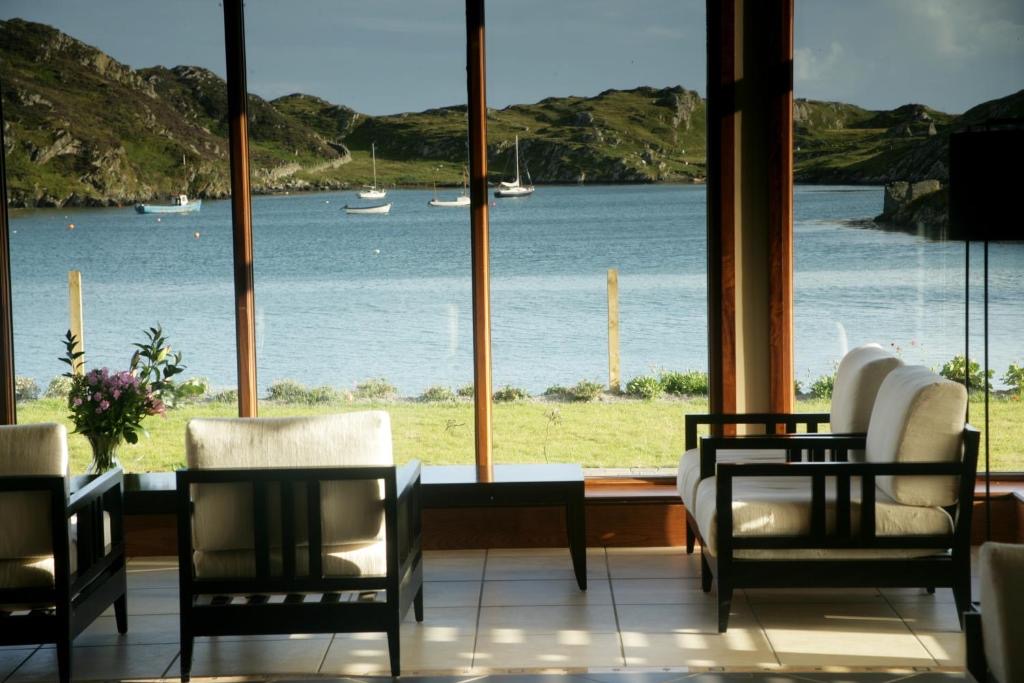 Zitgedeelte met witte stoelen met kussens en een kleine tafel met bloemen, met uitzicht op grote ramen. Het uitzicht buiten toont een kalme baai met zeilboten en rotsachtige, groene heuvels onder een bewolkte lucht - een idyllisch tafereel, perfect voor degenen die dromen van een vakantie in Ierland. | Echt Ierland Vakanties
