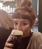 Een vrouw met bruin haar in een knot nipt van een pint Guinness, wat de geest van reisen Ierland belichaamt. Ze draagt een zwart topje met een lichtgrijs vest. De licht wazige pubsetting, flessen op planken en een andere persoon die gedeeltelijk zichtbaar is, roepen de charme op van een Iers uitje. | Echt Ierland Vakanties