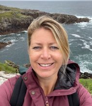 Een persoon die lacht op een selfie staat bij een ruige, rotsachtige kustlijn op hun rundreise Ierland. Ze dragen een kastanjebruine jas en hebben een zwarte rugzak bij zich. De golven van de oceaan beuken tegen de rotsen en op de achtergrond zijn grasachtige kliffen zichtbaar onder een grijze, bewolkte lucht. | Echt Ierland Vakanties