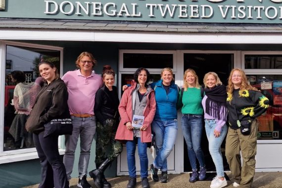 Een groep van negen mensen, genietend van hun rundreise Irland, staat buiten het Donegal Tweed Visitor Centre en lacht naar de camera. Casual gekleed, sommigen dragen jasjes tegen de Ierse wind. De groene gevel van het gebouw toont trots "Triona Donegal Tweed Visitor Centre" boven de ingang. | Echt Ierland Vakanties