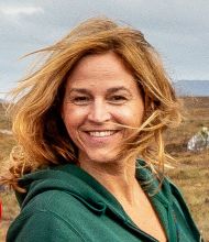 Een persoon met golvend, schouderlang blond haar lacht warm naar de camera, met een groene hoodie aan. De achtergrond hint naar een rondreis door Ierland, met grasvelden en verre heuvels onder een bewolkte lucht. Winderig haar suggereert een luchtige atmosfeer die ideaal is om de landschappen van Ierland te verkennen. | Echt Ierland Vakanties