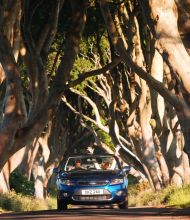 Een blauwe cabriolet rijdt over een weg met kronkelende, eeuwenoude bomen die een natuurlijke tunnel vormen op een betoverende rundreise Ierland. Twee passagiers zijn zichtbaar, een zwaait terwijl het zonlicht door de dichte takken filtert en gevlekte schaduwen werpt. Het tafereel straalt avontuur en natuurlijke schoonheid uit. | Echt Ierland Vakanties