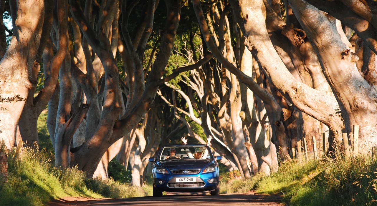 Een blauwe cabriolet rijdt over een weg met kronkelende, eeuwenoude bomen die een natuurlijke tunnel vormen op een betoverende rundreise Ierland. Twee passagiers zijn zichtbaar, een zwaait terwijl het zonlicht door de dichte takken filtert en gevlekte schaduwen werpt. Het tafereel straalt avontuur en natuurlijke schoonheid uit. | Echt Ierland Vakanties