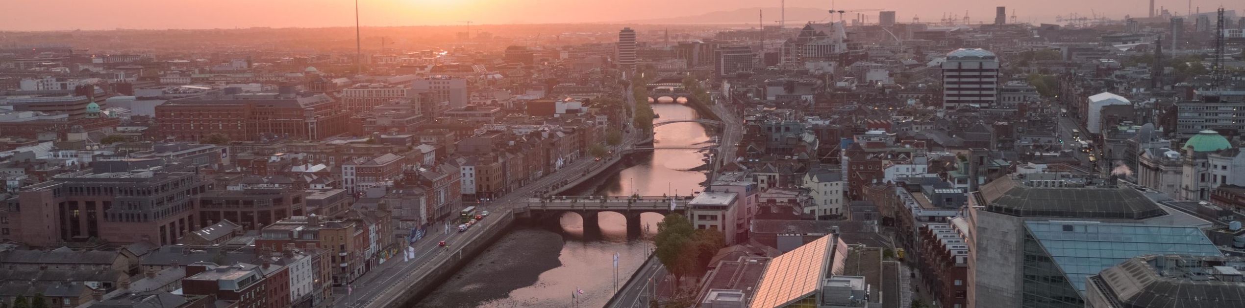Luchtfoto van Dublin bij zonsondergang, met de rivier de Liffey die door de stad slingert. Bruggen overspannen de rivier, met historische en moderne gebouwen aan de skyline. De Spire staat hoog, omarmd door een warme oranje lucht - een perfect tafereel voor degenen die een rondreis Ierland overwegen. | Echt Ierland Vakanties