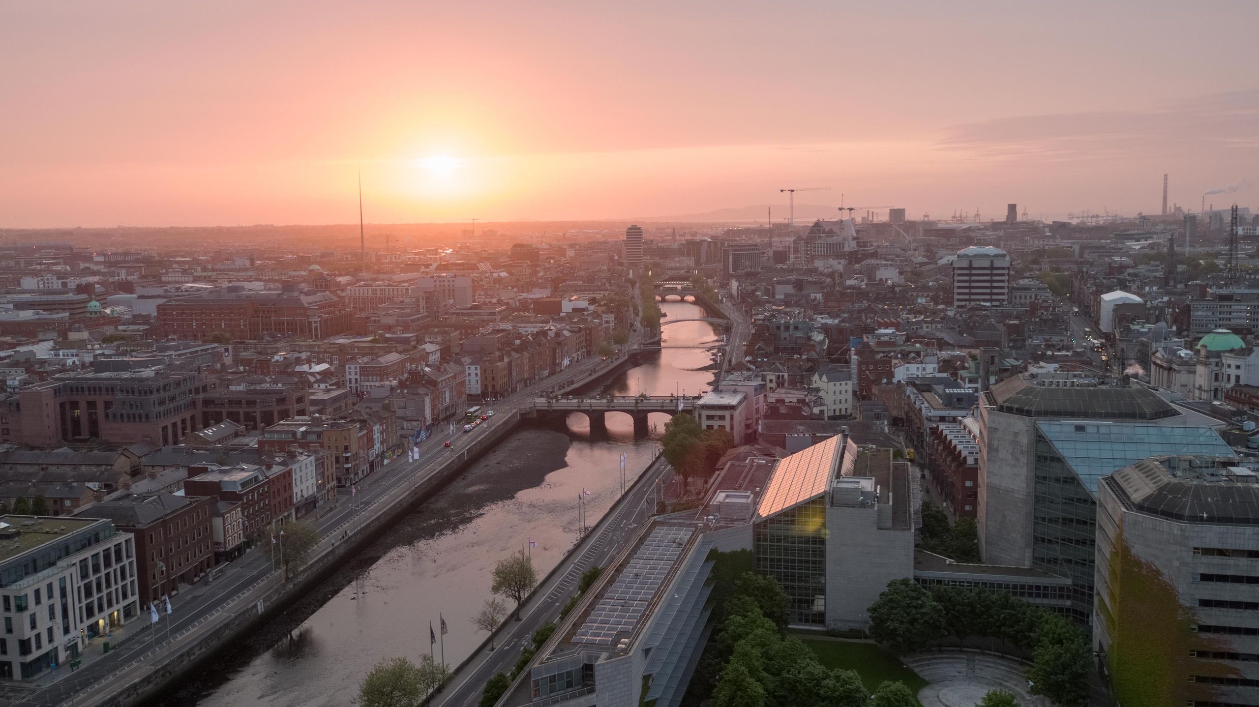 Luchtfoto van Dublin bij zonsondergang, met de rivier de Liffey die door de stad slingert. Bruggen overspannen de rivier, met historische en moderne gebouwen aan de skyline. De Spire staat hoog, omarmd door een warme oranje lucht - een perfect tafereel voor degenen die een rondreis Ierland overwegen. | Echt Ierland Vakanties