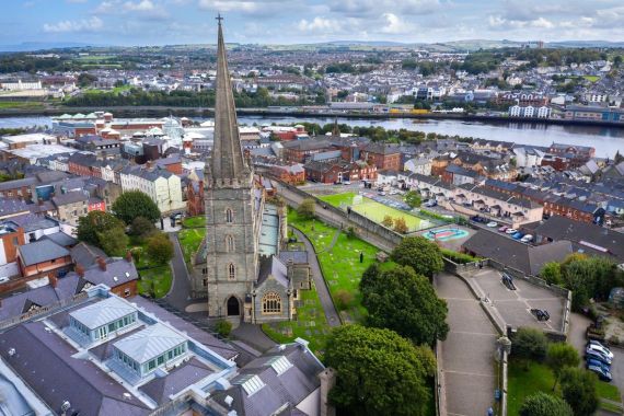 Luchtfoto van een stadsgezicht dat doet denken aan een rondreis Ierland, met een grote kerk met een hoge torenspits, omringd door weelderige groene gazons en bomen. Centraal gelegen tussen gebouwen en huizen, wordt het tafereel gecompleteerd door een rivier die erachter stroomt, met heuvels in de verte onder gedeeltelijk bewolkte luchten. | Echt Ierland Vakanties