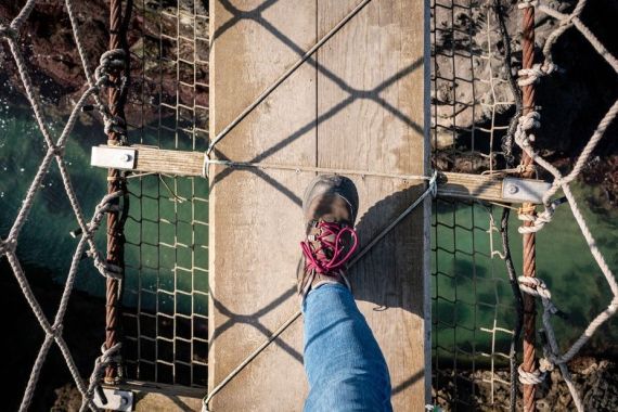 Een persoon loopt over een smalle touwbrug met een houten plankpad, en omarmt de sensatie van een vakantie in Ierland. Ze dragen blauwe jeans en bruine wandelschoenen met rode veters en zien hoe het licht schaduwen werpt die lijken op een net. Beneden stroomt water, wat bijdraagt aan het avontuur van reizen in Ierland. | Echt Ierland Vakanties