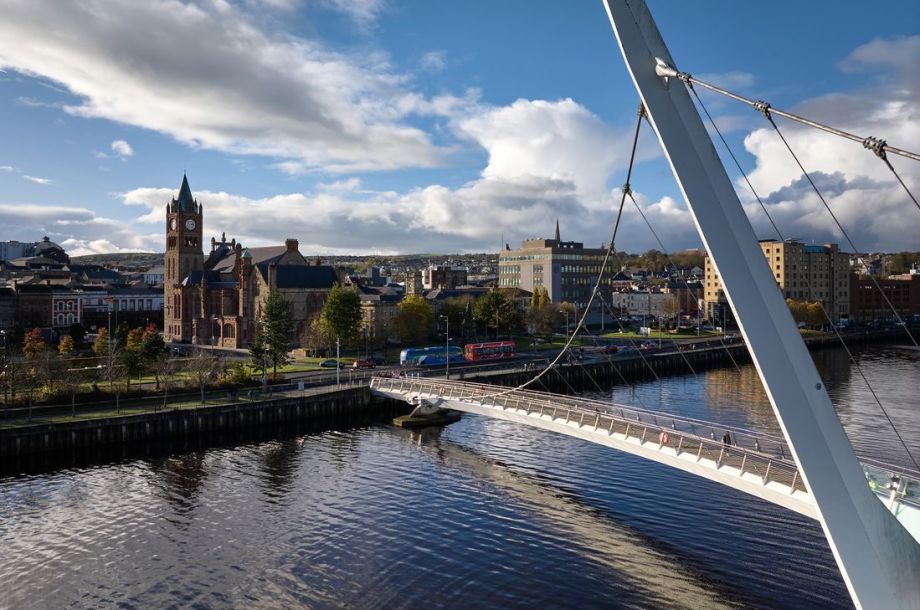 Uitzicht op een moderne witte brug met kabels over een kalme rivier in Ierland. Op de achtergrond een stadsgezicht met een prominente kerk met een hoge klokkentoren, omringd door verschillende gebouwen en bomen onder een gedeeltelijk bewolkte blauwe lucht - een ideaal tafereel voor elke reisen Ierland-route. | Echt Ierland Vakanties