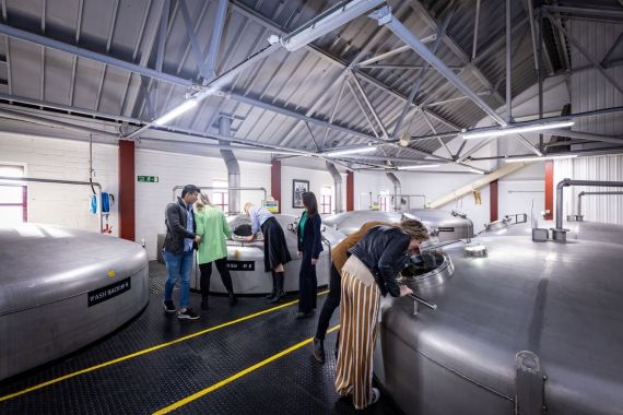Mensen bekijken grote metalen vaten in een industrieel gebouw met zichtbare balken en dakramen, waarschijnlijk op een rundreise Ierland. Bezoekers inspecteren de vaten, mogelijk tijdens een rondleiding door een brouwerij of distilleerderij. De vloer is gemarkeerd met gele lijnen en de kamer is goed verlicht. | Echt Ierland Vakanties
