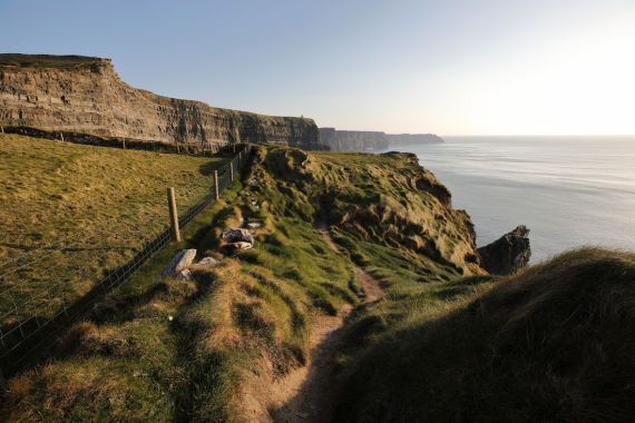 Een smal zandpad slingert langs de met gras begroeide klifwand, begrensd door een hek aan de linkerkant. De klif strekt zich uit in de verte onder een helderblauwe lucht. Beneden, terwijl u op uw rundreise irland verkent, strekt de kalme zee zich uit tot aan de horizon. Zonlicht werpt subtiele schaduwen, waardoor het ruige landschap wordt benadrukt. | Echt Ierland Vakanties