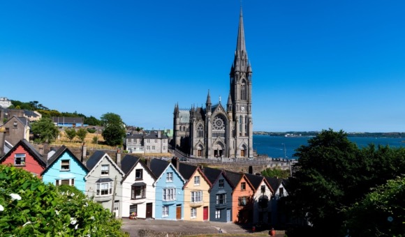 Een rij kleurrijke, schuine huizen in pastelblauw, geel, groen, roze en oranje kleuren siert de voorgrond. Dit pittoreske tafereel legt de essentie van een Urlaub in Ierland vast. Op de achtergrond staat een hoge kathedraal met een prominente torenspits, afgezet tegen een heldere lucht en weelderig groen aan het water. | Echt Ierland Vakanties