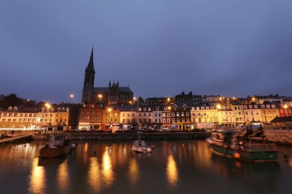 Een serene avondscène in een stad aan het water, perfect voor degenen die dromen van een vakantie in Ierland. Vage boten glijden op de voorgrond terwijl een kerk met een torenspits en kleurrijke gebouwen oplichten onder een schemerblauwe lucht; hun weerspiegelingen dansen op kalm water terwijl straatlantaarns een warme gloed werpen. | Echt Ierland Vakanties