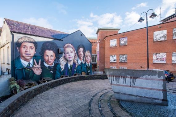 Een levendige muurschildering toont vijf schoolkinderen met verschillende gezichtsuitdrukkingen op een muur van een gebouw, wat de essentie van een urlaub in Ierland vastlegt. Het gebied bevat bakstenen gebouwen, een geplaveid pad en een stenen structuur met strepen. De gedeeltelijk bewolkte lucht werpt schaduwen bij de lantaarnpaal aan de rechterkant. | Echt Ierland Vakanties