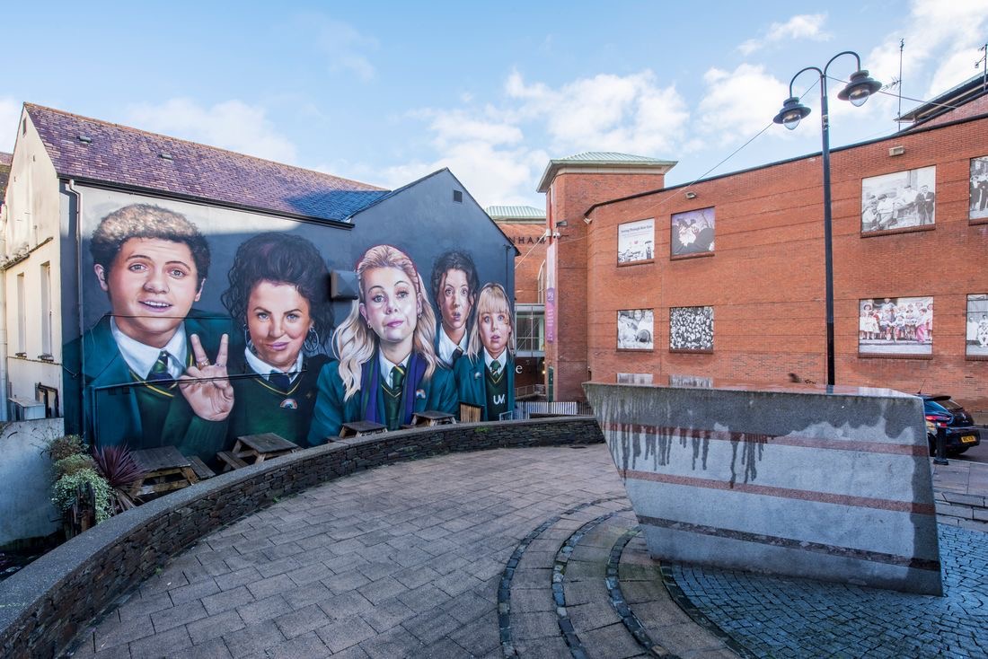 Een levendige muurschildering toont vijf schoolkinderen met verschillende gezichtsuitdrukkingen op een muur van een gebouw, wat de essentie van een urlaub in Ierland vastlegt. Het gebied bevat bakstenen gebouwen, een geplaveid pad en een stenen structuur met strepen. De gedeeltelijk bewolkte lucht werpt schaduwen bij de lantaarnpaal aan de rechterkant. | Echt Ierland Vakanties