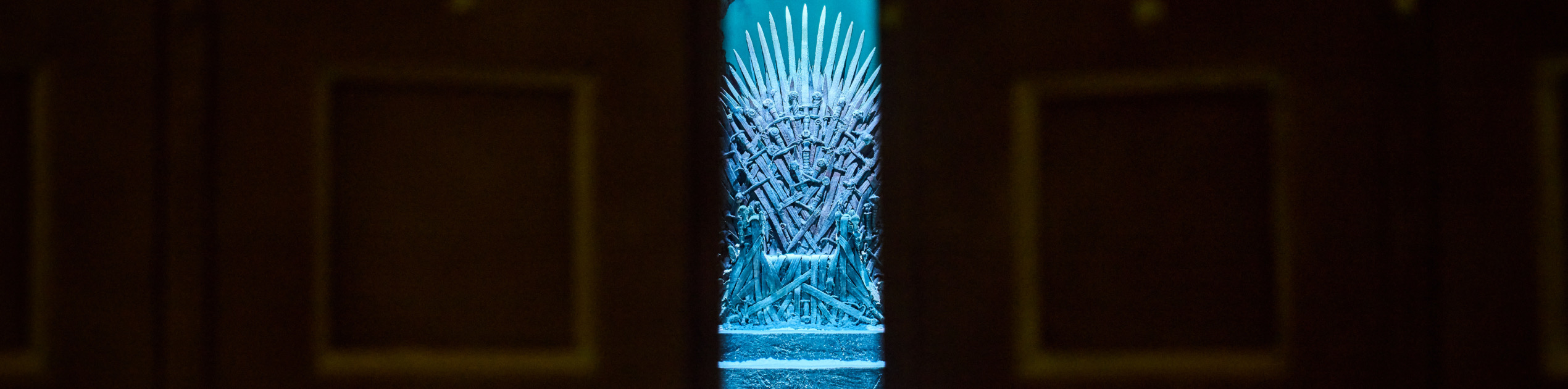 Een schemerige kamer met de Iron Throne in het midden van de achtergrond, bekeken door gedeeltelijk open, donkere houten dubbele deuren. De troon is gesilhouetteerd met een blauwe gloed, die doet denken aan oude kastelen die men zou kunnen verkennen tijdens een reizen Irland, rustend op stenen treden bestoven met sneeuw. | Echt Ierland Vakanties
