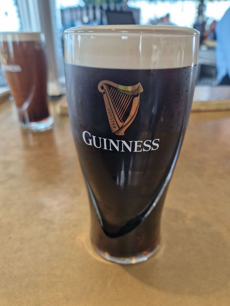 Een pintglas gevuld met donker Guinness-bier staat op een houten tafel, met het iconische logo met zijn harpontwerp. In de wazige achtergrond van een authentieke pub, die doet denken aan een rondreis Ierland, hint een tweede glas subtiel naar levendige verhalen en gedeeld gelach. | Echt Ierland Vakanties