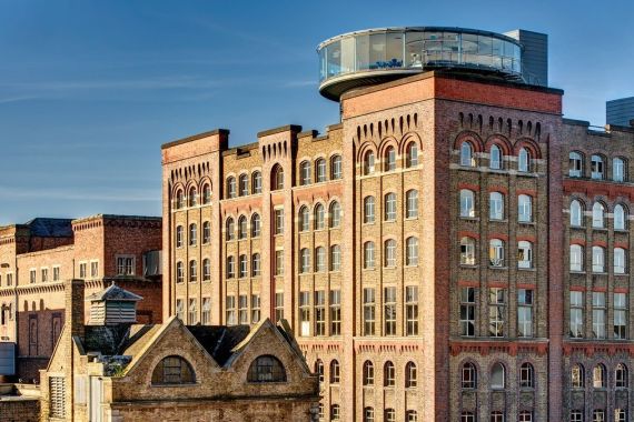 Een historisch gebouw van rode baksteen met boogramen en een rond glazen dak vangt een industriële maar toch eigentijdse sfeer, perfect voor iedereen die droomt van een "urlaub Irland"-ervaring. Met zijn bakstenen details en schoorstenen onder een helderblauwe lucht, combineert het modern design met klassieke charme. | Echt Ierland Vakanties