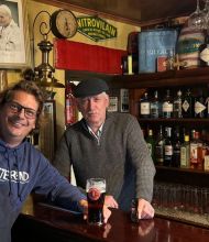 Twee mannen staan achter de bar van een gezellige pub in Ierland. De man links draagt een bril en een marineblauwe hoodie, lachend met een pint bier voor zich. De man rechts, met een platte pet en trui, leunt op de bar. Planken vol flessen drank en een vintage foto geven charme aan deze typisch Ierse stop op uw rundreise Irland. | Echt Ierland Vakanties