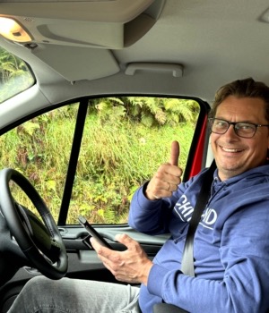 Een persoon met een bril, een blauwe hoodie en jeans, zit op de bestuurdersstoel van een rood voertuig. Ze lachen, steken hun duim omhoog en houden een telefoon vast, klaar om te beginnen aan hun "rondreis Ierland." Het raam onthult groen buiten, met varens zichtbaar op de achtergrond. | Echt Ierland Vakanties