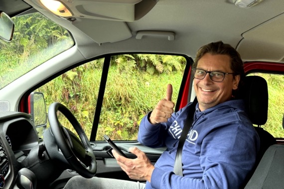 Een persoon met een bril, een blauwe hoodie en jeans, zit op de bestuurdersstoel van een rood voertuig. Ze lachen, steken hun duim omhoog en houden een telefoon vast, klaar om te beginnen aan hun "rondreis Ierland." Het raam onthult groen buiten, met varens zichtbaar op de achtergrond. | Echt Ierland Vakanties