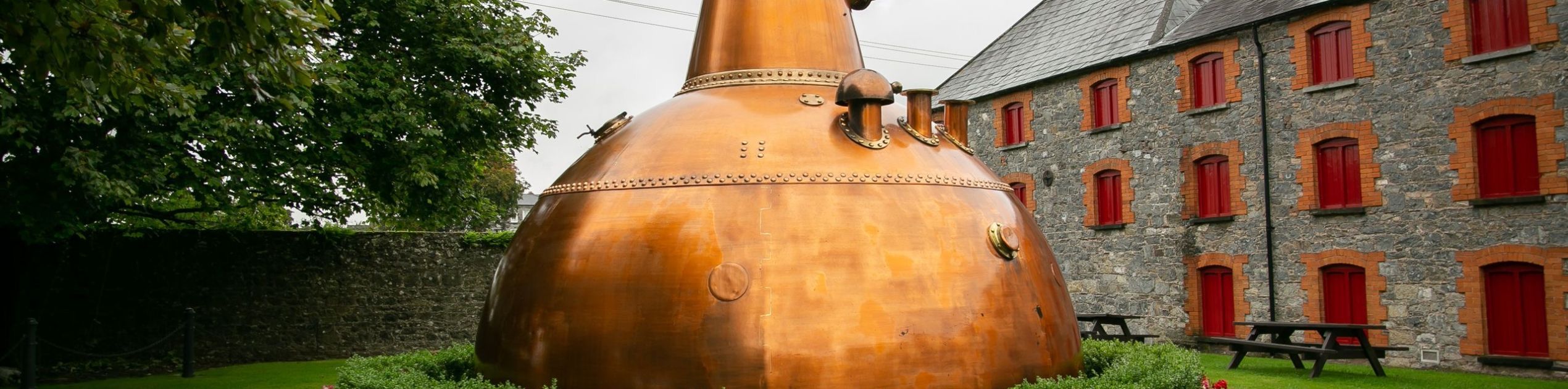 Buiten staat een grote koperen pot still, omringd door levendige bloeiende struiken en een stenen rand. Daarachter draagt een charmant gebouw bij aan de aantrekkingskracht van een rundreise Ierland, met zijn schuine dak en rode luiken. Een bewolkte lucht torent hoog boven ons uit, terwijl het gebladerte van de bomen het tafereel links omlijst. | Echt Ierland Vakanties