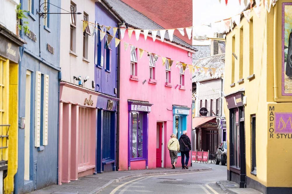 Een charmant stedelijk tafereel dat doet denken aan een rondreis Ierland, met levendige gebouwen in tinten blauw, roze en geel. Twee mensen lopen over de smalle weg terwijl er boven hen kleurrijke vlaggen wapperen, die de schilderachtige en levendige essentie van deze betoverende omgeving vastleggen. | Echt Ierland Vakanties