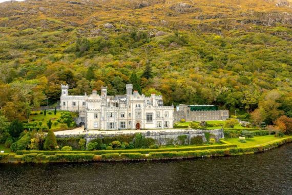 Een groots, historisch kasteel met grijze stenen muren en talloze torentjes ligt te midden van weelderig groen in een scène die doet denken aan een vakantie in Ierland. Gelegen aan een sereen meer, met beboste bergen erachter, is het landschap rijk aan herfstkleuren, wat de pittoreske schoonheid versterkt. | Echt Ierland Vakanties