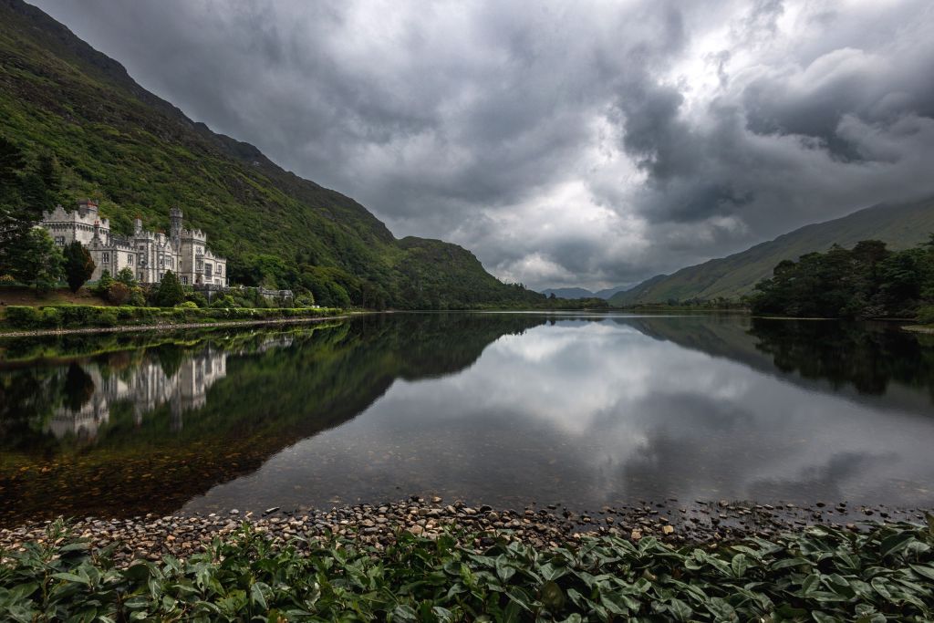 Kasteel aan een meer onder een bewolkte lucht in betoverend Ierland. Het gebouw staat links, omgeven door dicht groen. Het stille water weerspiegelt dramatische wolken en weelderige bergen, terwijl een kiezelstrand en bladplanten de voorgrond omlijsten. Perfect voor vakanties in Ierland met zijn kalme, sfeervolle atmosfeer. | Echt Ierland Vakanties