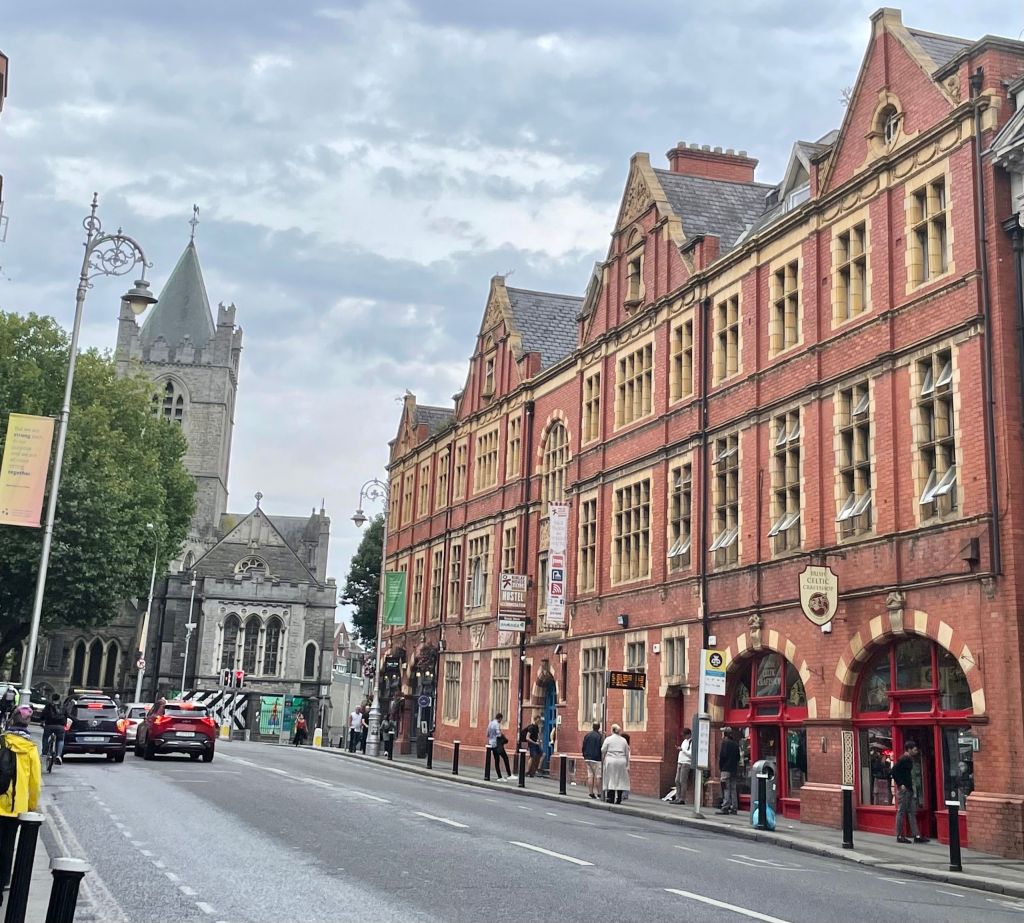 Straatbeeld met historische bakstenen gebouwen aan de rechterkant, met boogramen en deuropeningen. Mensen slenteren er langs, verzonken in hun rundreise Ierland. Een stenen kerktoren is zichtbaar op de achtergrond. Meerdere auto's rijden over de weg onder een bewolkte lucht bij bomen aan de linkerkant. | Echt Ierland Vakanties